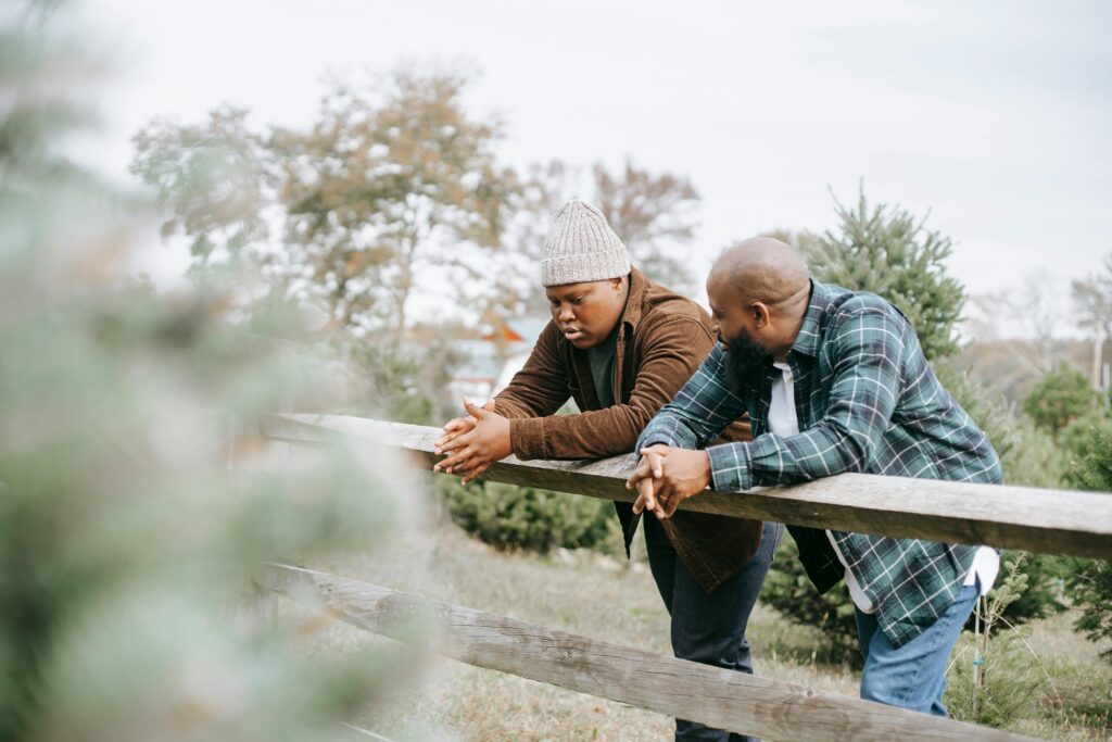 Two black men having a conversation while leaning on wooden fence in wooded area